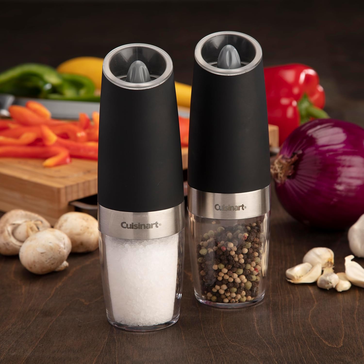 Shows the salt and pepper grinders in a real kitchen setting, surrounded by fresh ingredients, to demonstrate their practical use in food preparation.
