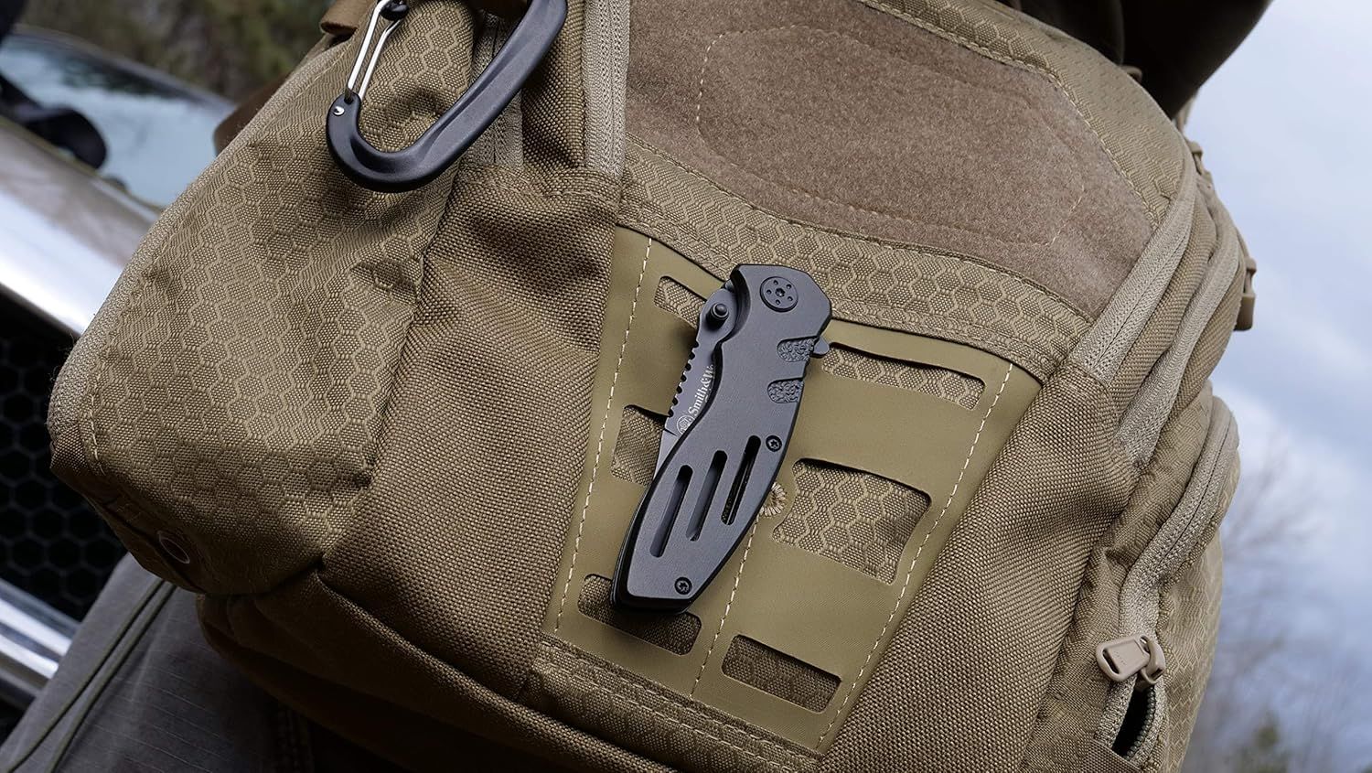 Demonstrates the product in a real-world tactical/outdoor context, showing how the multi-tool knife can be conveniently attached to gear via carabiner for easy access during outdoor activities.