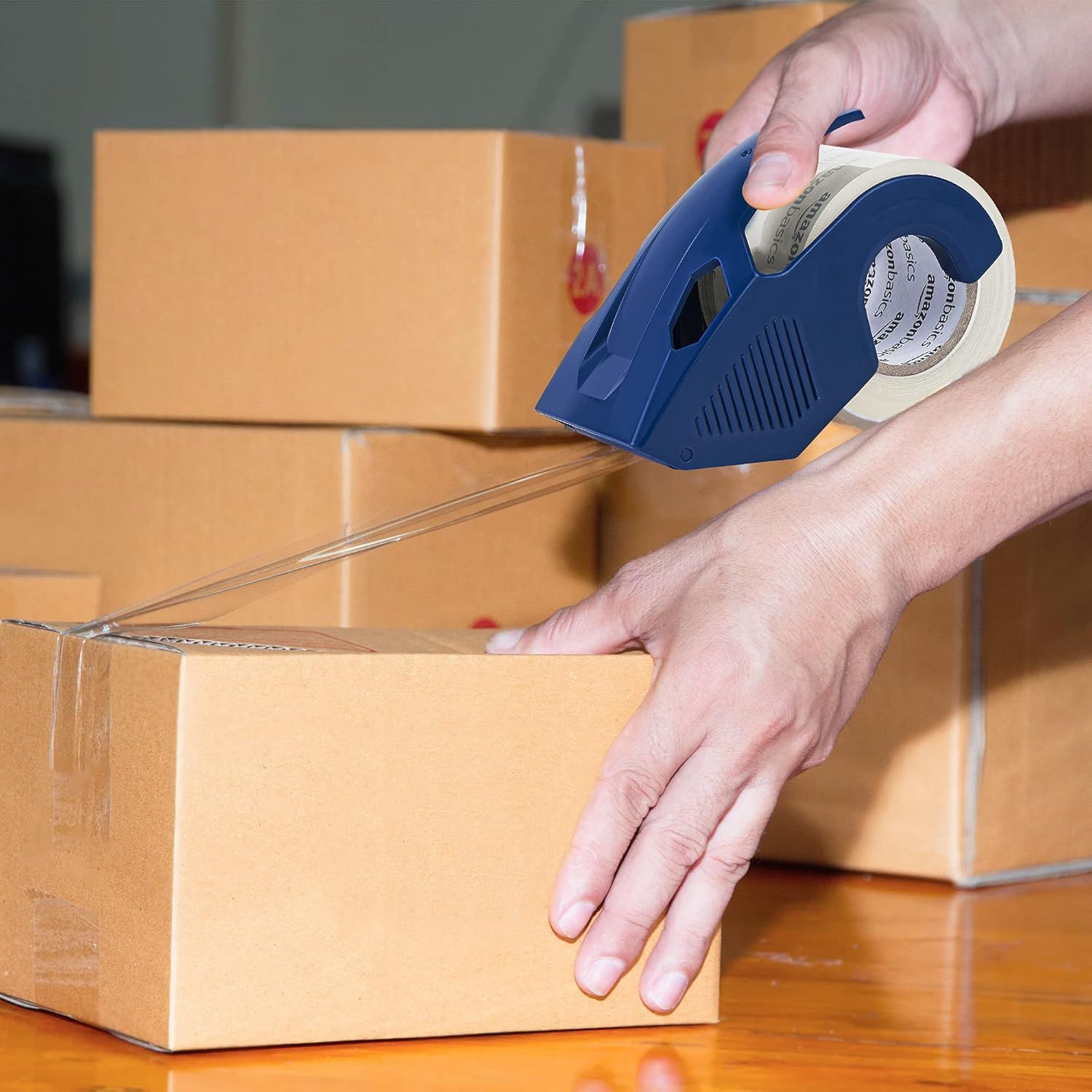 Demonstrates the product in a real-world context, showing someone using the tape dispenser to seal cardboard boxes, helping shoppers visualize practical everyday use.