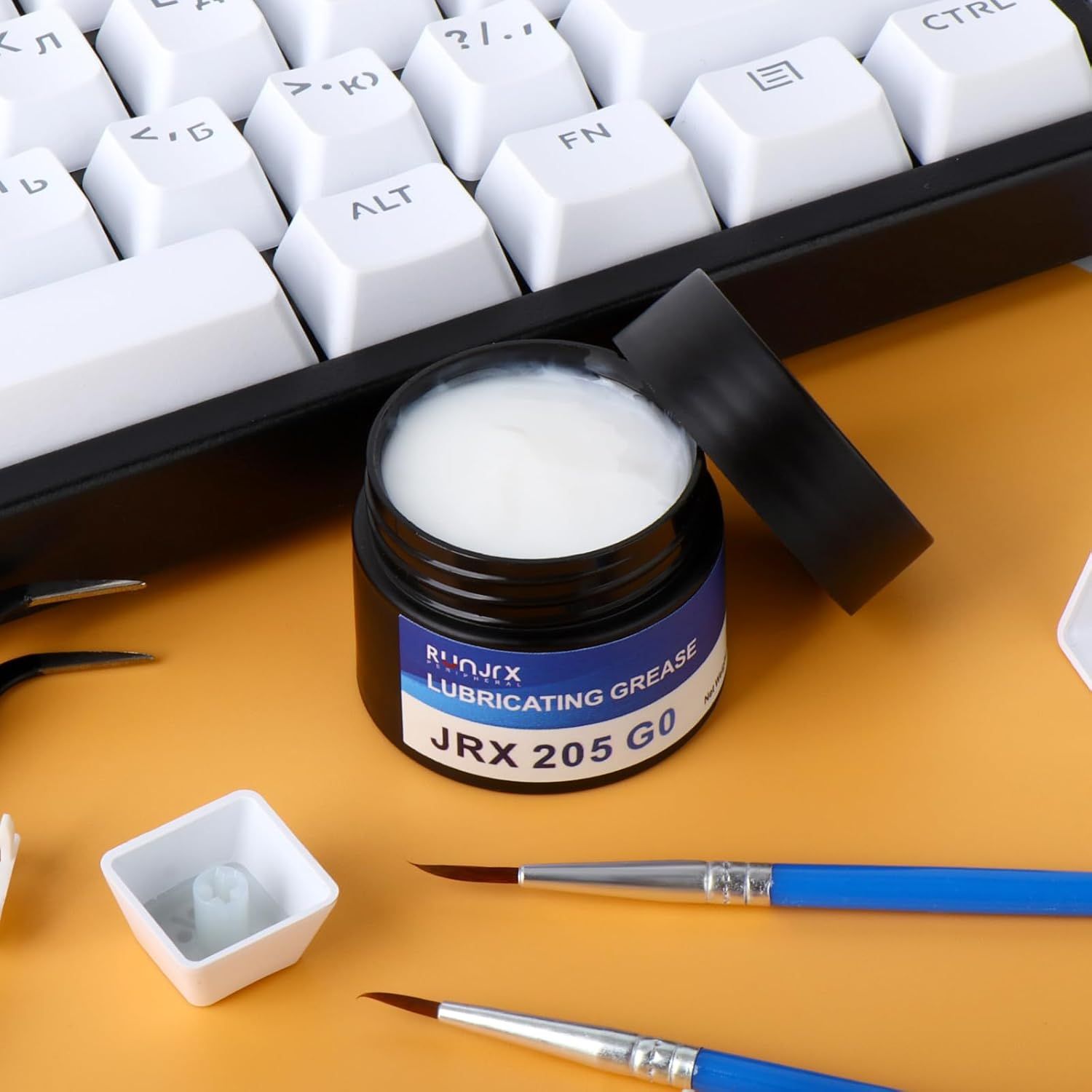 Displays the lubricating grease product alongside keyboard switches and application tools to show the product in its intended use context for mechanical keyboard maintenance.