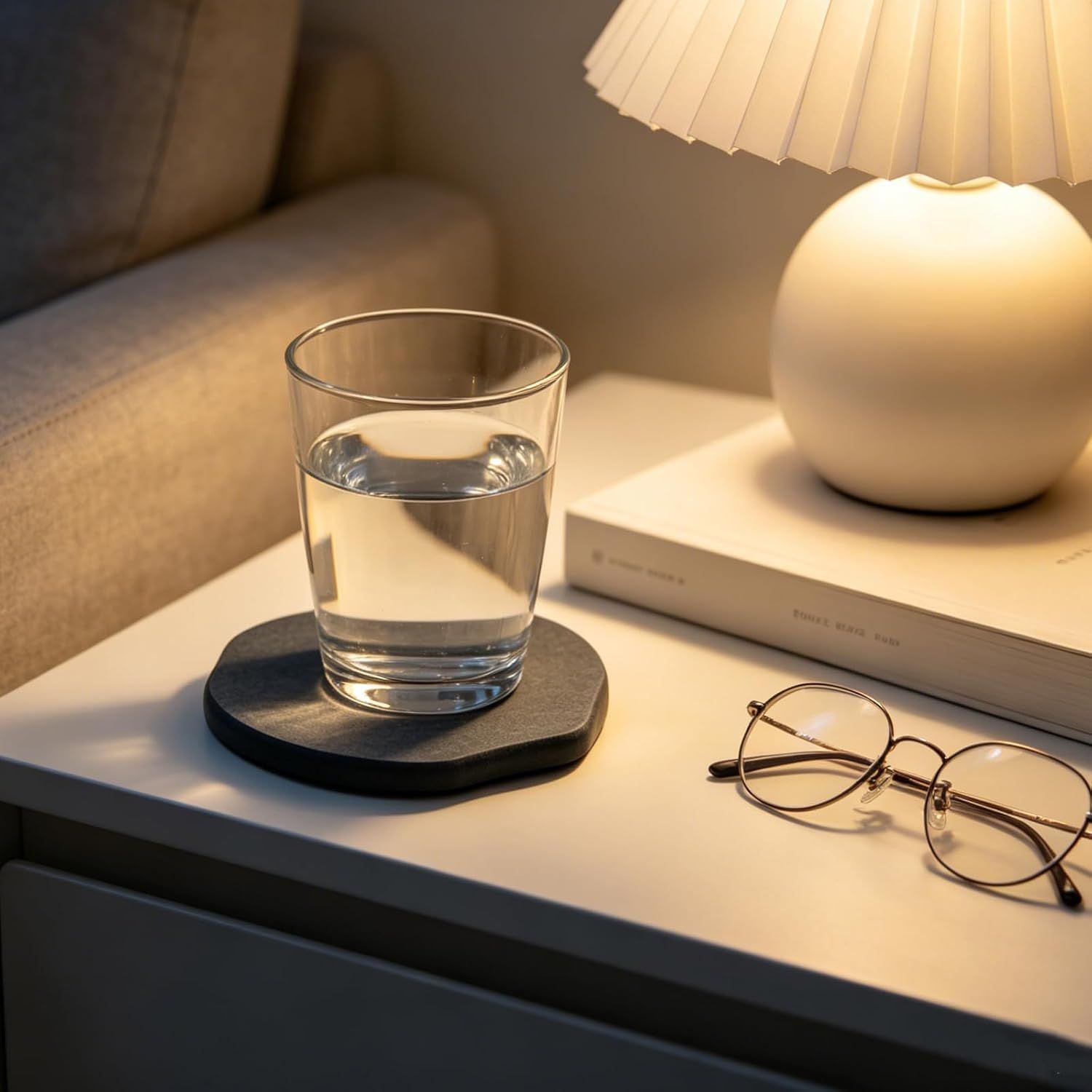 Shows the coaster in a bedroom nightstand setting with a glass of water, demonstrating its versatility across different rooms in the home.